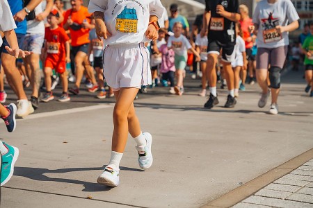 Kinder laufen bei einem Schülerlauf auf einer abgesperrten Strecke; im Vordergrund läuft ein Kind in weißer Sportkleidung.