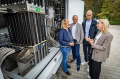 Vier Personen stehen neben dem Elektrolyseur und sprechen vor der technischen Anlage mit Rohren, Leitungen und Behältern.