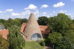 Historischer kegelförmiger Tafelturm der Glashütte Gernheim mit Backsteinfassade und großem Fenster, umgeben von roten Dächern und Bäumen.