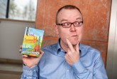 Mann mit Brille und blauem Hemd hält eine Packung des OWL-Heimat-Quiz in der Hand und legt nachdenklich einen Finger ans Kinn.
