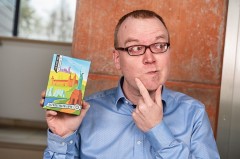 Mann mit Brille und blauem Hemd hält eine Packung des OWL-Heimat-Quiz in der Hand und legt nachdenklich einen Finger ans Kinn.