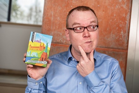 Mann mit Brille und blauem Hemd hält eine Packung des OWL-Heimat-Quiz in der Hand und legt nachdenklich einen Finger ans Kinn.
