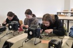 Drei junge Personen arbeiten an Lötstationen in einem Seminarraum.
