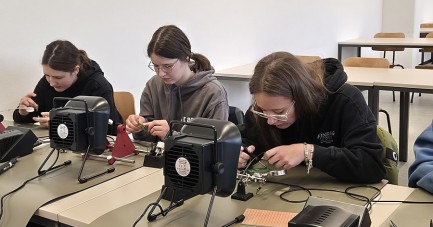 Drei junge Personen arbeiten an Lötstationen in einem Seminarraum.