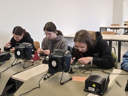 Drei junge Personen arbeiten an Lötstationen in einem Seminarraum.
