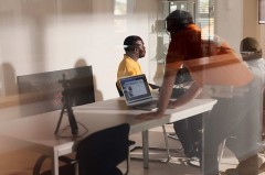 Drei Personen arbeiten in einem hellen Technikraum an einem Tisch mit Laptop und technischer Ausrüstung; die Szene ist durch eine Glasscheibe aufgenommen.