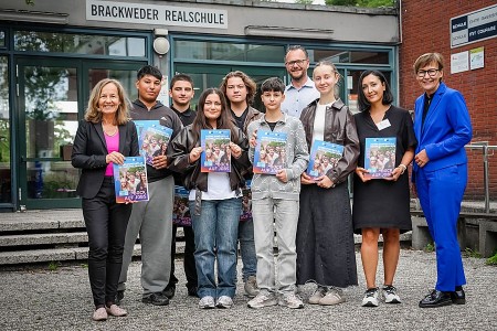 chülerinnen und Schüler der Brackweder Realschule halten gemeinsam mit Erwachsenen Exemplare des IHK-Ausbildungsatlas 2027 vor dem Schulgebäude.