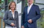 Eine Frau im grauen Blazer und ein Mann im dunkelblauen Anzug stehen nebeneinander vor einer großen Glasfassade.