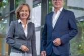 Eine Frau im grauen Blazer und ein Mann im dunkelblauen Anzug stehen nebeneinander vor einer großen Glasfassade.
