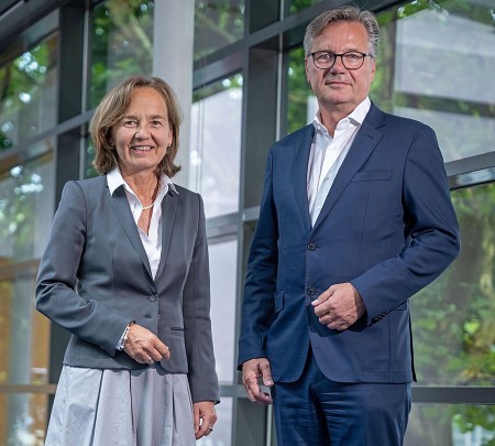 Eine Frau im grauen Blazer und ein Mann im dunkelblauen Anzug stehen nebeneinander vor einer großen Glasfassade.