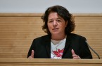 Eine Frau spricht an einem Rednerpult und gestikuliert vor einer hellen Holzvertäfelung.