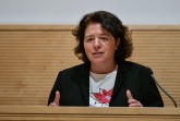 Eine Frau spricht an einem Rednerpult und gestikuliert vor einer hellen Holzvertäfelung.