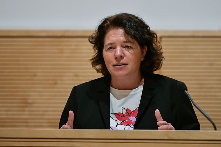 Eine Frau spricht an einem Rednerpult und gestikuliert vor einer hellen Holzvertäfelung.