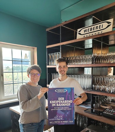 Zwei Personen halten im Weberei-Bahnhof ein Plakat mit der Aufschrift „Der Kneipenabend im Bahnhof“.