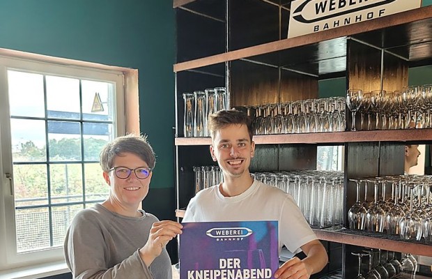 Zwei Personen halten im Weberei-Bahnhof ein Plakat mit der Aufschrift „Der Kneipenabend im Bahnhof“.