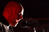 Sänger bei einem Konzert, der vor dunklem Hintergrund in ein Mikrofon singt und von rotem Licht beleuchtet wird.