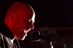 Sänger bei einem Konzert, der vor dunklem Hintergrund in ein Mikrofon singt und von rotem Licht beleuchtet wird.