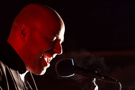 Sänger bei einem Konzert, der vor dunklem Hintergrund in ein Mikrofon singt und von rotem Licht beleuchtet wird.