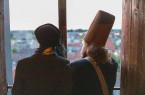 Zwei von hinten fotografierte Personen mit historischen Kopfbedeckungen blicken aus einem Fenster auf die Stadt.