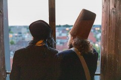 Zwei von hinten fotografierte Personen mit historischen Kopfbedeckungen blicken aus einem Fenster auf die Stadt.
