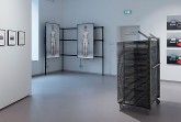 Ausstellungsraum mit gerahmten Fotografien, zwei Bildschirmen mit menschlichen Figuren und einer Metallinstallation.