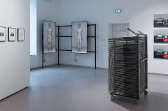 Ausstellungsraum mit gerahmten Fotografien, zwei Bildschirmen mit menschlichen Figuren und einer Metallinstallation.