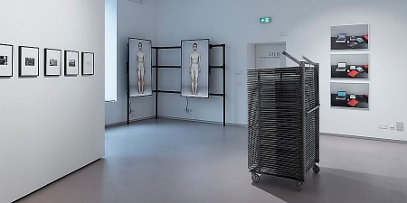Ausstellungsraum mit gerahmten Fotografien, zwei Bildschirmen mit menschlichen Figuren und einer Metallinstallation.