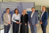 Fünf Personen bei der Eröffnung der Riga-Ausstellung im Foyer der Bezirksregierung Detmold vor einer Ausstellungstafel.