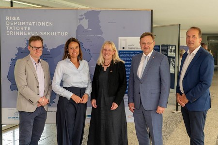 Fünf Personen bei der Eröffnung der Riga-Ausstellung im Foyer der Bezirksregierung Detmold vor einer Ausstellungstafel.