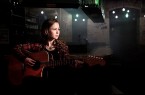Sandrine Lisken sitzt mit einer Akustikgitarre in einem dunkel beleuchteten Raum und blickt zur Seite.