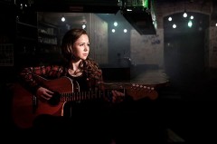 Sandrine Lisken sitzt mit einer Akustikgitarre in einem dunkel beleuchteten Raum und blickt zur Seite.