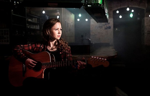 Sandrine Lisken sitzt mit einer Akustikgitarre in einem dunkel beleuchteten Raum und blickt zur Seite.
