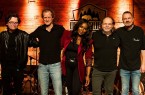 Fünf Mitglieder einer Band stehen nebeneinander auf einer Bühne vor einer orange beleuchteten Wand.