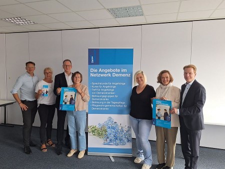 Sieben Beteiligte von DeineDiakonie und der Sparkassenstiftung stehen neben einem Banner mit Angeboten des Netzwerks Demenz und halten Veranstaltungsplakate.